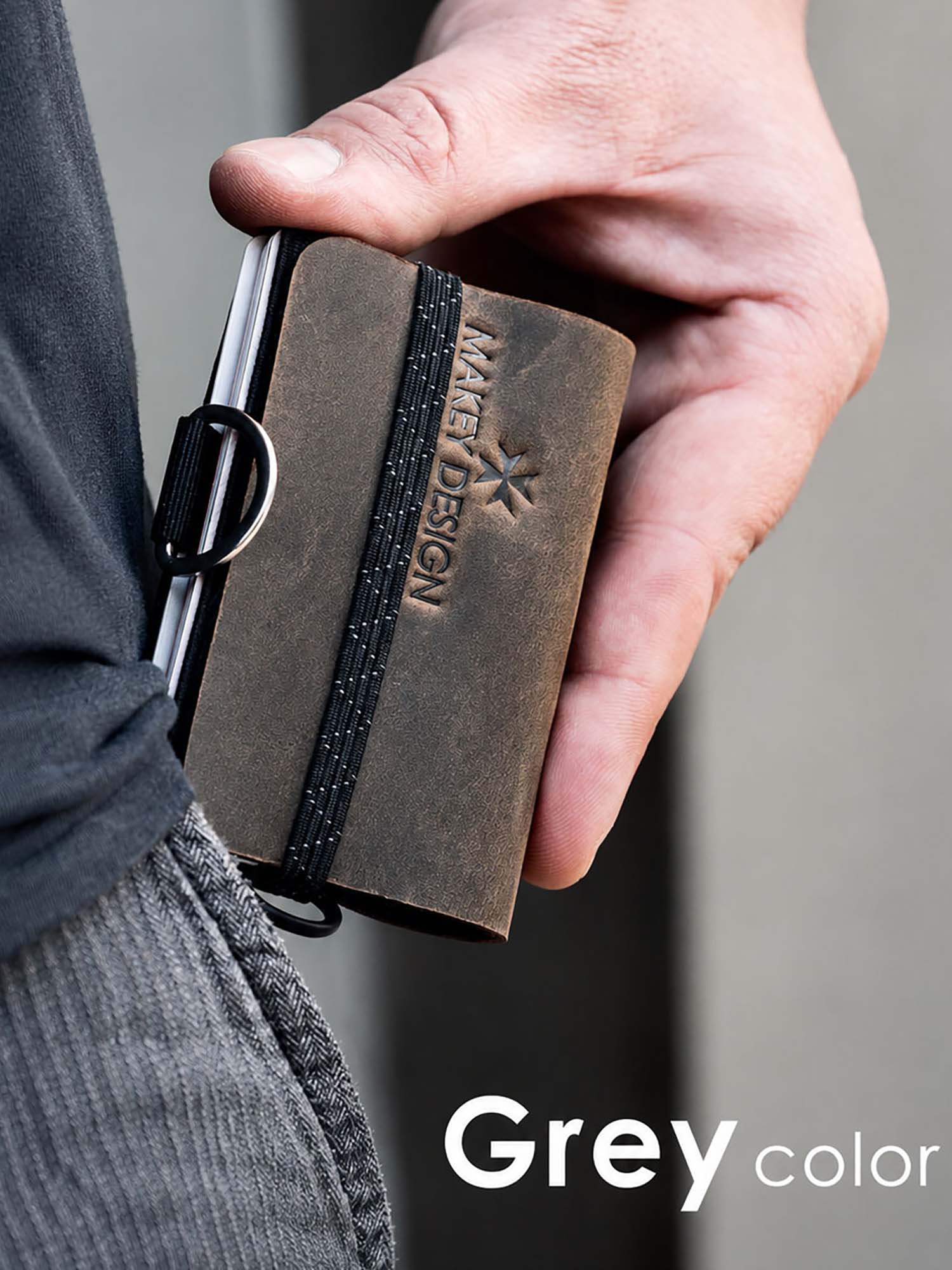 Hand holding a brown leather wallet with 'MAKEY DESIGN' branding against a gray background.